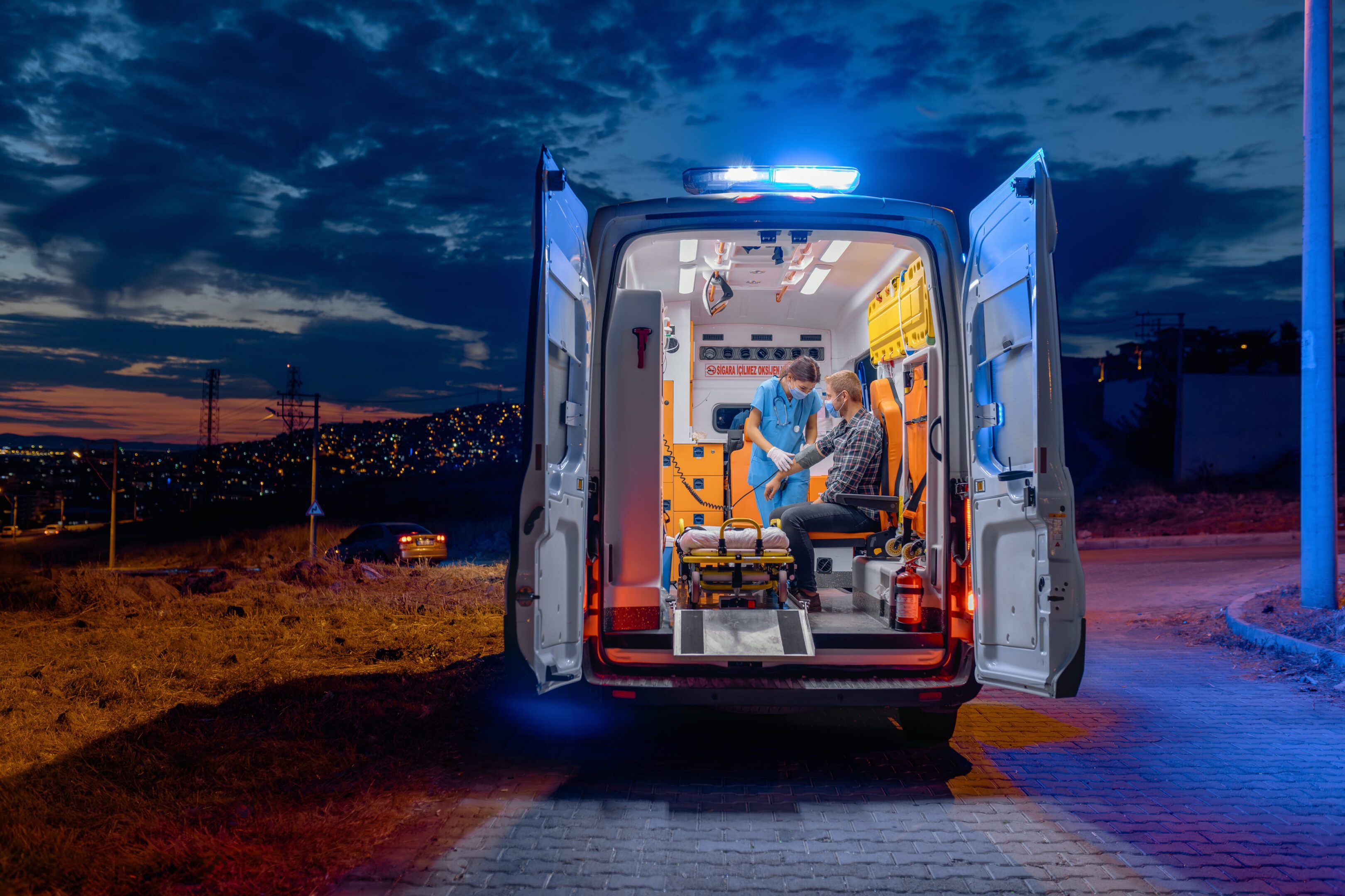 A first aider healthcare emergency staff nurse woman wearing a medical face mask taking care of a man patient sitting on stretcher chair taking and measuring patients blood pressure inside a medical service ambulance car at night